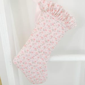 Könnte beinhalten: Ein hellrosa Weihnachtsstrumpf mit einem sich wiederholenden Muster aus kleinen rosa Rosen und grünen Blättern. Der Strumpf hat oben einen gerüschten Stoffaufschlag und eine passende hellrosa Bandschlaufe zum Aufhängen.