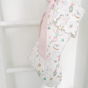 May include: A white quilted Christmas stocking featuring a repeating pattern of Nutcracker-themed illustrations including dancers, rocking horses, and bells. The stocking has a ruffled cuff and a long, light pink ribbon bow attached at the top.
