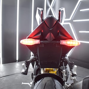 May include: Rear view of a black motorcycle with illuminated red taillights and turn signals. The motorcycle has a sleek design, with the brand name "Aprilia" visible on the license plate holder. The image is taken indoors with bright, linear lighting.