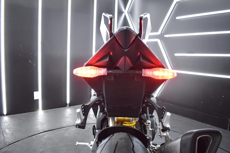 May include: Rear view of a black motorcycle with illuminated red taillights and turn signals. The motorcycle has a sleek design, with the brand name "Aprilia" visible on the license plate holder. The image is taken indoors with bright, linear lighting.