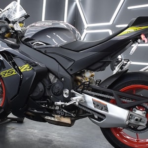 May include: A black and gray Aprilia RS 660 motorcycle with red wheels and yellow accents. The motorcycle is parked indoors and has a sleek, aerodynamic design. The wheels are a striking red color, and the bike has the text "RS 660" in yellow.