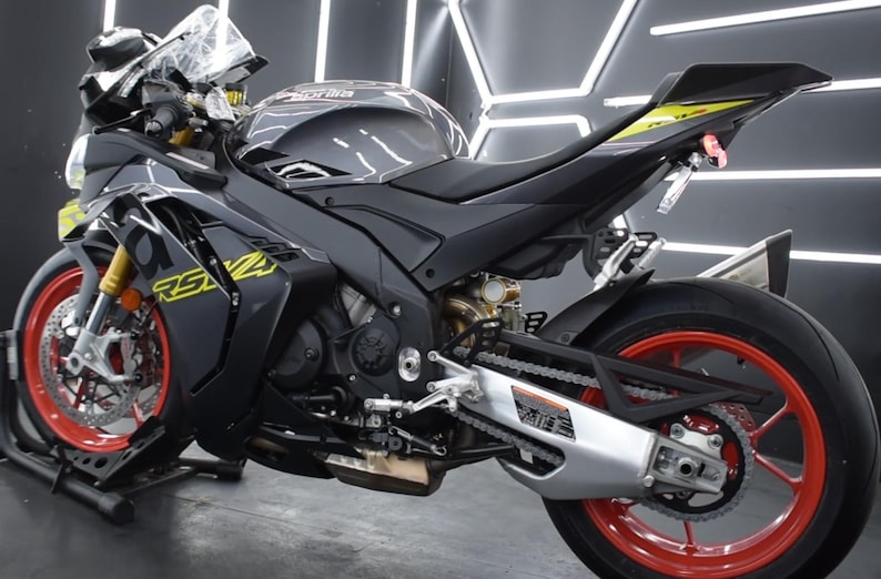 May include: A black and gray Aprilia RS 660 motorcycle with red wheels and yellow accents. The motorcycle is parked indoors and has a sleek, aerodynamic design. The wheels are a striking red color, and the bike has the text "RS 660" in yellow.