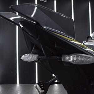 May include: Close-up of a black motorcycle tail section with clear turn signals. The motorcycle has a sleek, modern design with a glossy finish. The background features geometric lighting.
