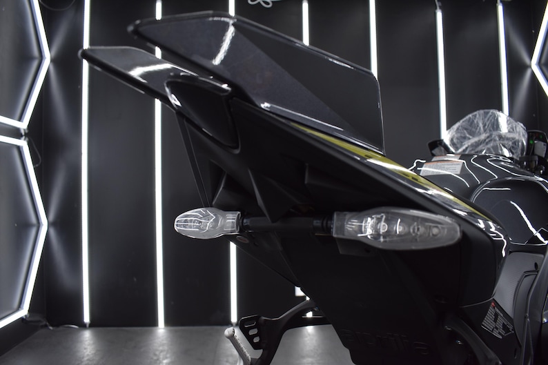 May include: Close-up of a black motorcycle tail section with clear turn signals. The motorcycle has a sleek, modern design with a glossy finish. The background features geometric lighting.