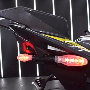 May include: Close-up of a black motorcycle tail section with a yellow accent stripe. The tail lights are illuminated red, and the turn signals are amber. The motorcycle is in a showroom setting.
