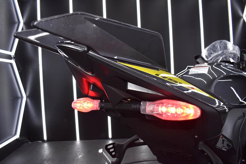 May include: Close-up of a black motorcycle tail section with a yellow accent stripe. The tail lights are illuminated red, and the turn signals are amber. The motorcycle is in a showroom setting.