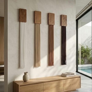 May include: Four vertical wall hangings, each featuring a rectangular wooden top piece with long, straight hanging cords in white, beige, light brown, and dark brown, arranged in a row above a low, light-toned wooden storage console unit.