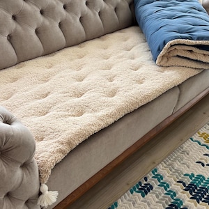 May include: A beige tufted sofa cushion with a blue and white throw blanket on top. The sofa cushion is made of a soft, fluffy material and has a textured surface.