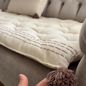 Könnte beinhalten: Ein weißes Kissen mit braunen Nähten und einem braunen Quasten. Das Kissen liegt auf einem grauen Sofa.