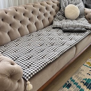 May include: A black and white gingham sofa cushion with tassels. The cushion is placed on a brown sofa with a tufted back.