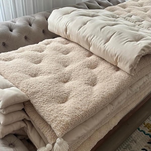 Könnte beinhalten: Ein Stapel beiger, getufteter und strukturierter Decken und Kissen. Die Decken haben eine flauschige, sherpaähnliche Textur und sind ordentlich gefaltet. Die Kissen sind glatt und rechteckig. Das Thema ist gemütliche Wohnkultur.