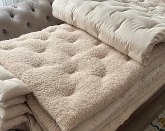 May include: A stack of beige, tufted, and textured blankets and cushions. The blankets have a plush, sherpa-like texture and are folded neatly. The cushions are smooth and rectangular. The scene suggests a cozy, comfortable home decor theme.