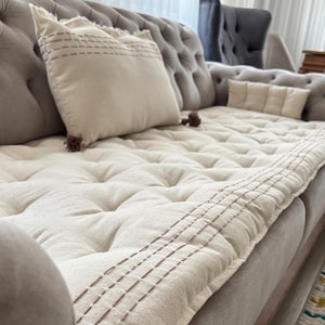 Puede incluir: Un cojín de sofá crema y cojines a juego. El cojín tiene un borde cosido y está colocado sobre un sofá gris. También hay dos cojines cuadrados y un pequeño cojín rectangular en el sofá. Los cojines tienen costuras decorativas y borlas.