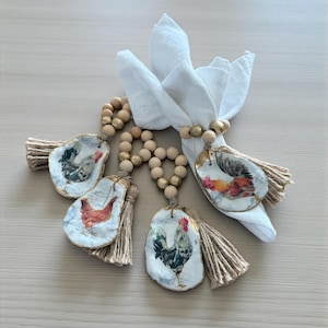 May include: A collection of decorative napkin rings featuring rooster illustrations. Each ring is crafted from a natural oyster shell, adorned with a rooster image, and accented with a jute tassel and wooden beads. A white napkin is folded in the center.