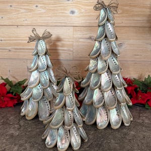 May include: Three decorative Christmas trees made of iridescent shells. Each tree is cone-shaped, with a twine bow at the top. The shells are arranged to resemble evergreen branches. Red poinsettias are visible in the background.