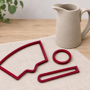 May include: A matte beige ceramic pitcher with a handle and spout, accompanied by three dark red geometric shapes: a large trapezoidal frame with a curved side, a small circular ring, and a long, narrow rectangular frame with rounded ends.