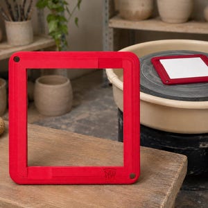 May include: A square red pottery bat system frame with rounded corners and a small circular hole in one corner. The frame features a subtle embossed logo in the bottom right corner. A matching smaller red square frame is visible on a pottery wheel.