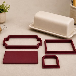 May include: A cream-coloured ceramic butter dish with a lid, accompanied by four dark red plastic cookie cutters in various rectangular and arched shapes, arranged on a flat surface alongside other kitchen items.