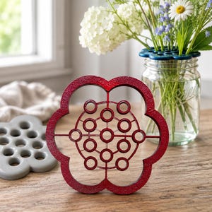 May include: A flower-shaped red metal flower frog with a grid of circular openings. The object features a textured finish and a central geometric pattern of interconnected rings. A similar gray flower frog sits nearby on the wooden surface.