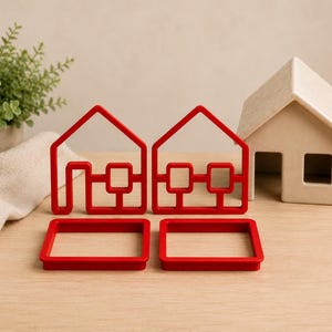 May include: Two red house-shaped cookie cutters with matching red rectangular frames, positioned in front of a small, light-colored, solid house-shaped object. The red cutters feature a simple outline of a house with a door and window grid design.