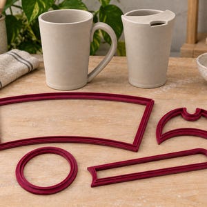 May include: A set of four dark red plastic clay cutters in various shapes, including a large curved trapezoid, a crescent moon shape, a small circle, and a long narrow rectangle with rounded ends, arranged on a flat wooden surface.