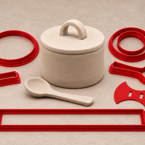 May include: A beige ceramic lidded jar and matching spoon surrounded by various red plastic cookie cutters, including a large rectangle, a circle, a small rectangle, a spoon-shaped cutter, and a curved piece with two rectangular slots.