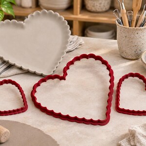 May include: A light gray heart-shaped tray with a scalloped edge and three red heart-shaped cookie cutters of varying sizes, each featuring a scalloped border design. The items are arranged on a flat surface alongside a wooden rolling pin.