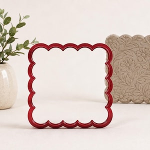 May include: A red square cookie cutter with a scalloped edge. The cutter has a hollow centre and a smooth, glossy finish. It is positioned upright, showcasing its distinct wavy perimeter and four-sided shape, designed for cutting dough or clay.