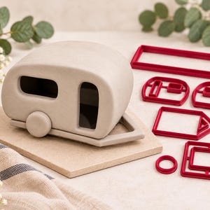 May include: A light gray camper trailer figurine with a smooth finish, featuring a rounded roof, a circular wheel, and rectangular window and door cutouts, accompanied by a set of six red plastic geometric cookie cutters of various shapes.