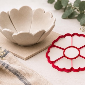 Peut inclure: Un bol en céramique beige avec un bord festonné en forme de pétales et un emporte-pièce en plastique rouge assorti. Le bol présente une finition mate lisse, tandis que l'emporte-pièce comporte un cercle central entouré de six segments.