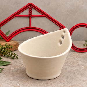 May include: A matte off-white ceramic bowl with a slanted rim and three small circular holes on one side, accompanied by a red plastic arch-shaped cutter and a red plastic circular cutter, all arranged on a neutral stone-textured surface.