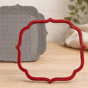 May include: A red quatrefoil-shaped cookie cutter positioned in the foreground. Behind it sits a gray square-shaped tile or coaster featuring a repeating embossed leaf pattern with rounded corners and decorative points on each side.