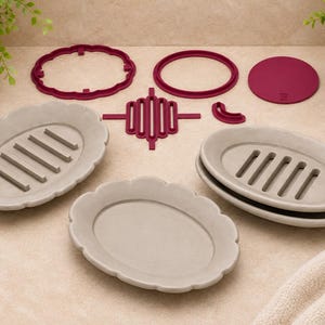 May include: A collection of three oval soap dishes in a light beige color, featuring scalloped edges and internal drainage slats. Accompanying the dishes are several dark red plastic components, including two frames, a solid disc, and a grid insert.