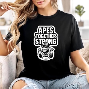 May include: Black t-shirt with a white graphic that reads "APES TOGETHER STRONG." The graphic includes a cartoon ape wearing sunglasses and holding money. The phrase "TO THE MOON" is also included.