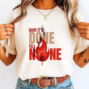 May include: Cream-colored t-shirt with the text "Until It's Done Tell No One" in red and brown. A stylized red flame graphic with a matchstick is centered below the text. The shirt is made of a soft material.