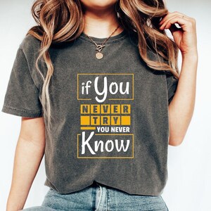 May include: Dark gray t-shirt with the text "if You Never Try You Never Know" in a yellow and white design. The shirt has short sleeves and a crew neck. The model is wearing a gold necklace.