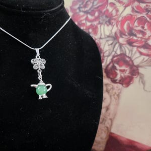 Könnte beinhalten: Eine silberfarbene Halskette mit einem schmetterlingsförmigen Anhänger und einem kleinen Teekannen-Anhänger mit einem grünen Edelstein. Die Halskette wird auf einer schwarzen Samtbüste präsentiert, mit einem Blumengemälde im Hintergrund.