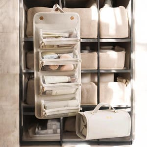 May include: A set of two beige travel makeup bags. The larger bag has a hanging loop and clear compartments to hold cosmetics. The smaller bag has a handle and the initials "D.A." embroidered on the front.