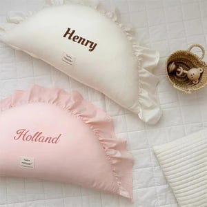 May include: Two half-moon shaped pillows with ruffled edges. The white pillow has the name "Henry" embroidered on it. The pink pillow has the name "Holland" embroidered on it.
