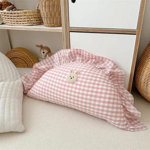 May include: A pink and white gingham pillow with a ruffled edge and a small embroidered bunny face in the center.