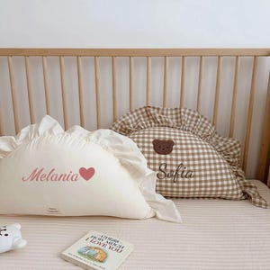 May include: Two white and brown gingham pillows with ruffled edges. One pillow has the name "Melania" embroidered on it with a pink heart. The other pillow has the name "Sofia" embroidered on it with a brown bear applique.