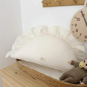 May include: A white ruffled pillow with a cream-colored label on a wicker basket. A brown and white polka dot doll with a pacifier is lying on the pillow.