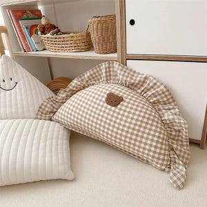 May include: A brown and white gingham pillow with a ruffled edge and a brown embroidered bear face. The pillow is on a white surface.