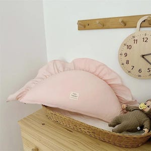 May include: A pink half-moon shaped pillow with ruffled edges. The pillow is sitting on a wicker basket.