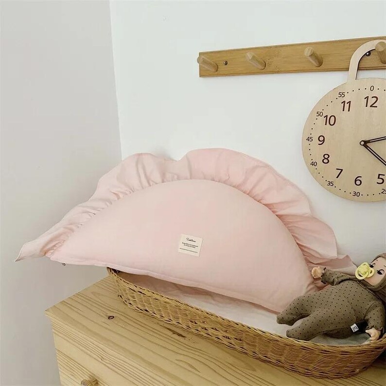 May include: A pink half-moon shaped pillow with ruffled edges. The pillow is sitting on a wicker basket.