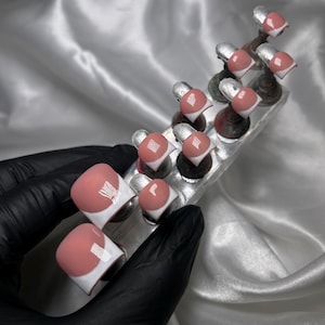 May include: A set of press-on toenails with a white and pink design. The nails are square-shaped with a white border and a pink center. The nails are displayed on a clear plastic holder.