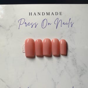 May include: A set of five glossy, peach-colored press-on nails with a square-rounded shape, displayed on a white marble-patterned card featuring the text HANDMADE Press On Nails and thank you for your order in dark blue script lettering.