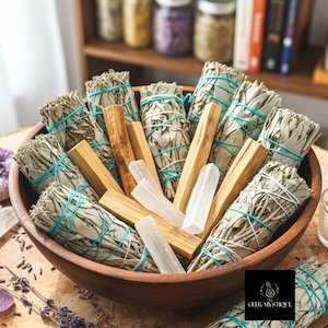 May include: A wooden bowl filled with bundles of white sage, palo santo sticks, and selenite crystals. The sage bundles are tied with blue twine. The bowl sits on a wooden surface, with jars and books in the background.