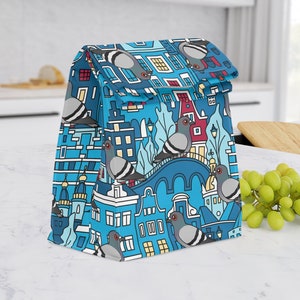 May include: A blue lunch bag featuring a whimsical illustration of a cityscape with pigeons. The design includes buildings, bridges, and trees in shades of blue, red, and yellow. The bag is sitting on a white countertop.