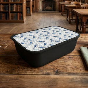 May include: A black rectangular lunchbox with a patterned lid featuring blue dachshunds on a white background. The lunchbox is sitting on a wooden table. A green napkin and a ceramic cup are also on the table.